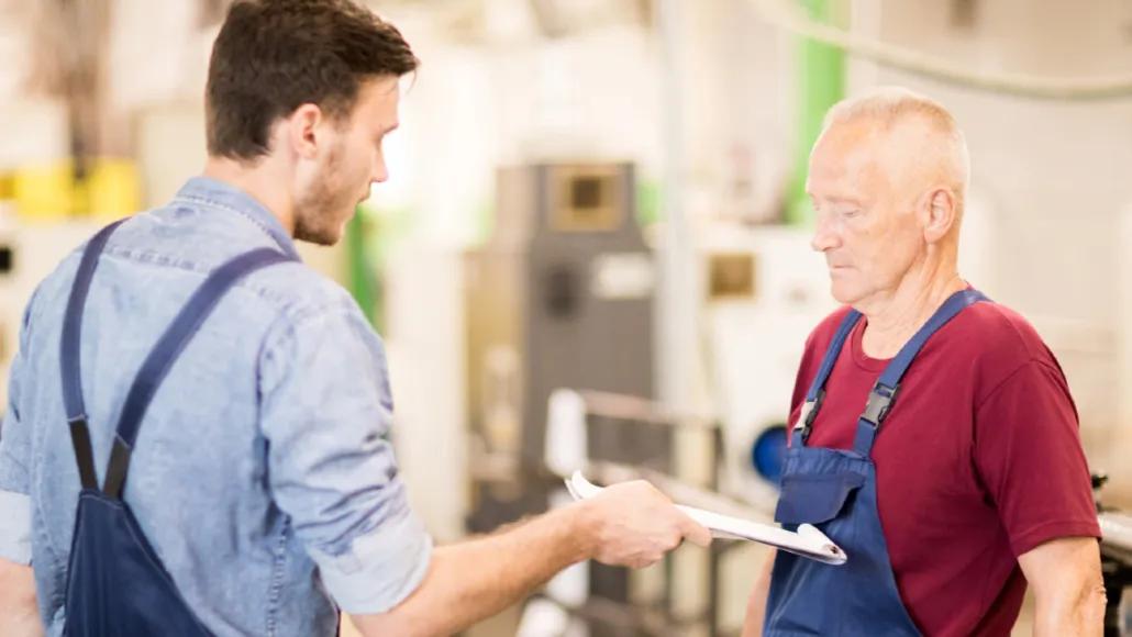 A young worker in overalls thrusts a clipboard with paperwork at an older person. The older person, also in overalls, is looking down and resting their left hand on a workbench behind them.