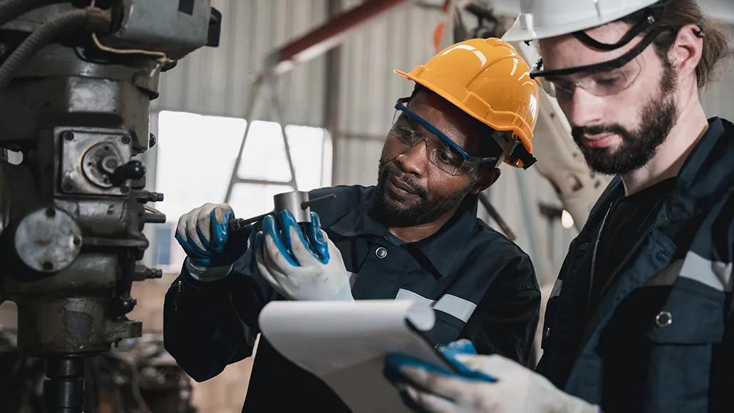 Two blue-collar workers in helmets, safety goggles, and gloves work on machinery.