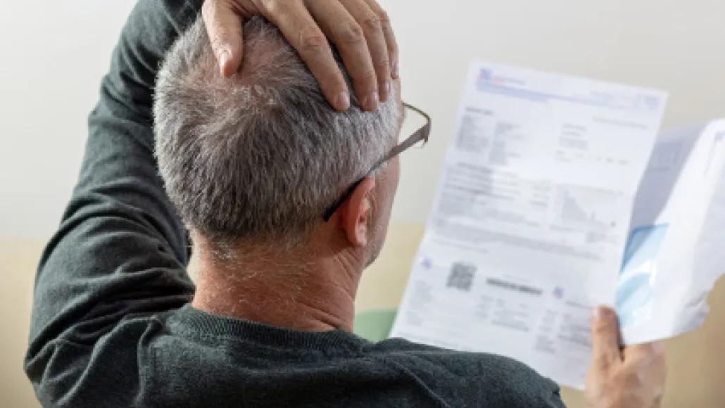 An older man with glasses has his left hand on top of his head while his right hand holds an open bill.