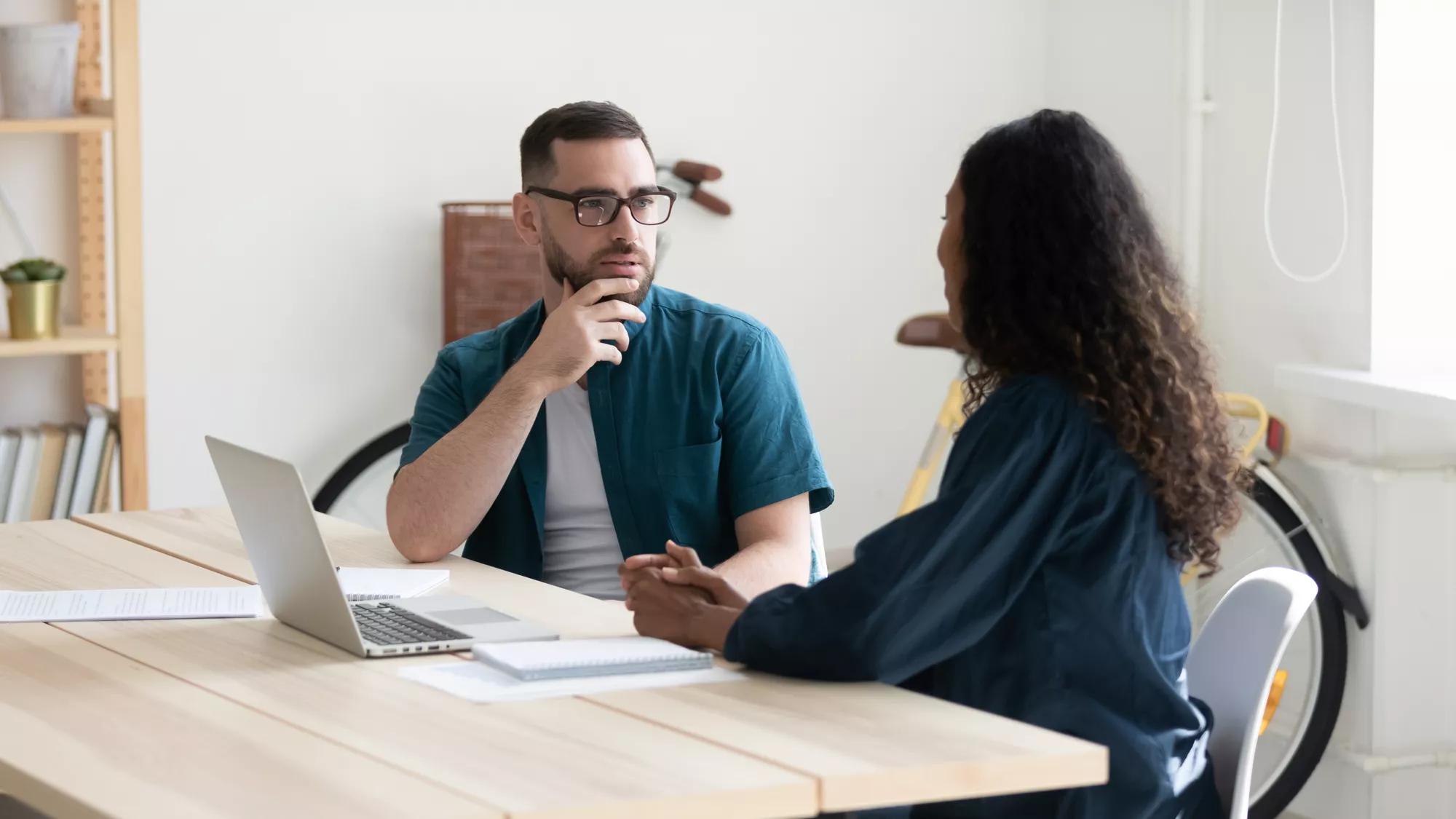 Thoughtful diverse colleagues sit at desk in office talk discuss business ideas at briefing together, pensive businesspeople brainstorm cooperate using laptop at meeting, collaboration concept