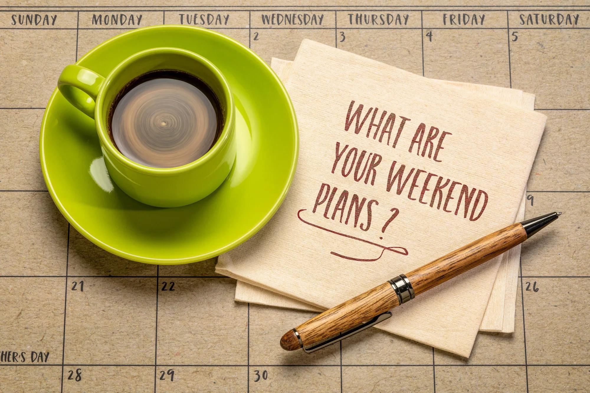 A cup of coffee, a pen, and two napkins on top of a calendar month. "What are your weekend plans?" is printed on the top napkin.