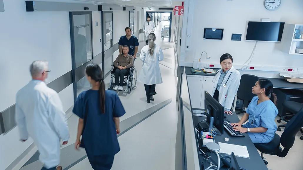 A photograph shows various hospital workers walking through a hospital's hallway and seated at the nurse's station.