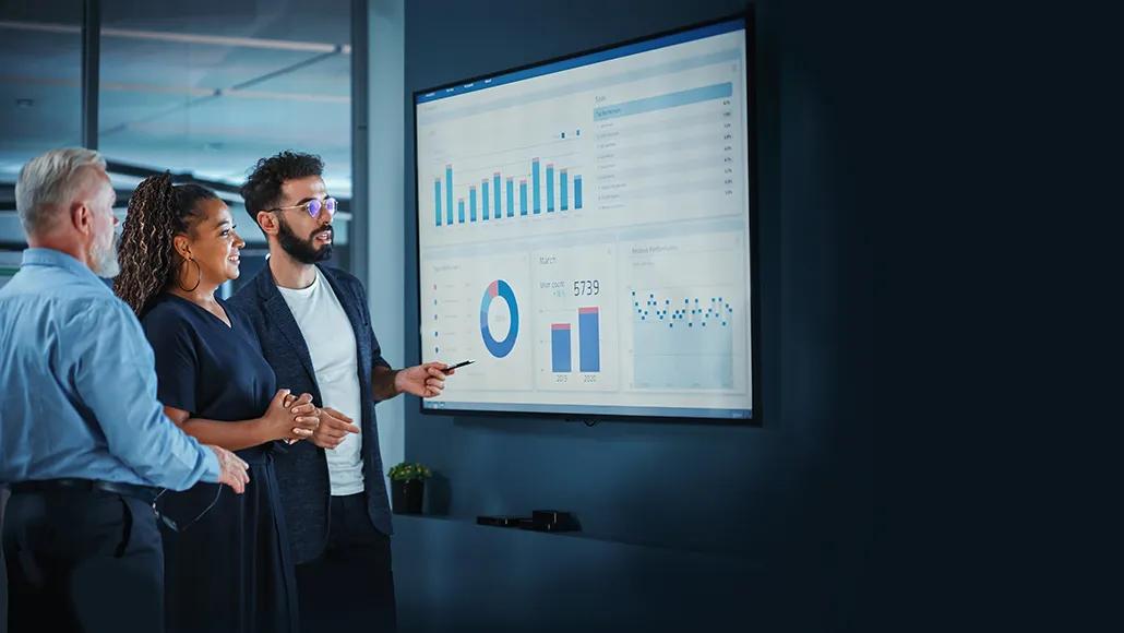 Three people consider a dashboard together. The dashboard has a bar graph, pie chart, bar chart, dot plot, and table.