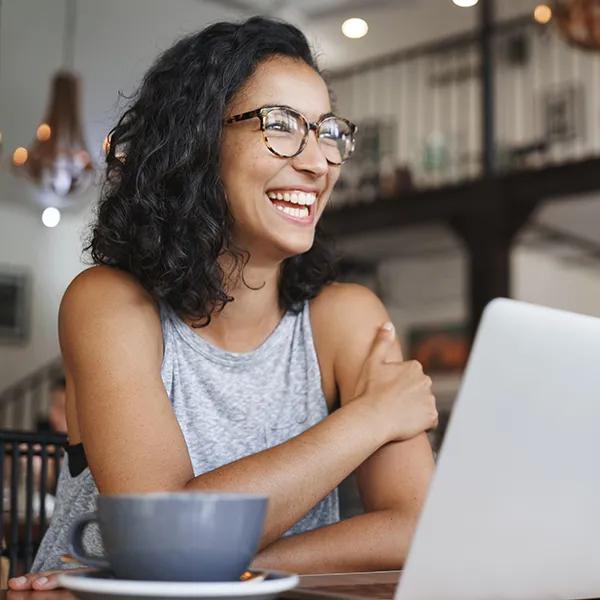 A smiling young woman sits in a cafe with a cup of coffee working on her laptop.