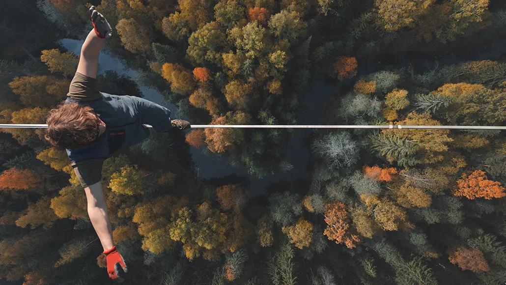 A bird's-eye shot of a person in a T-shirt, jumpsuit, and climbing gloves walking a highwire with a forest and river below.