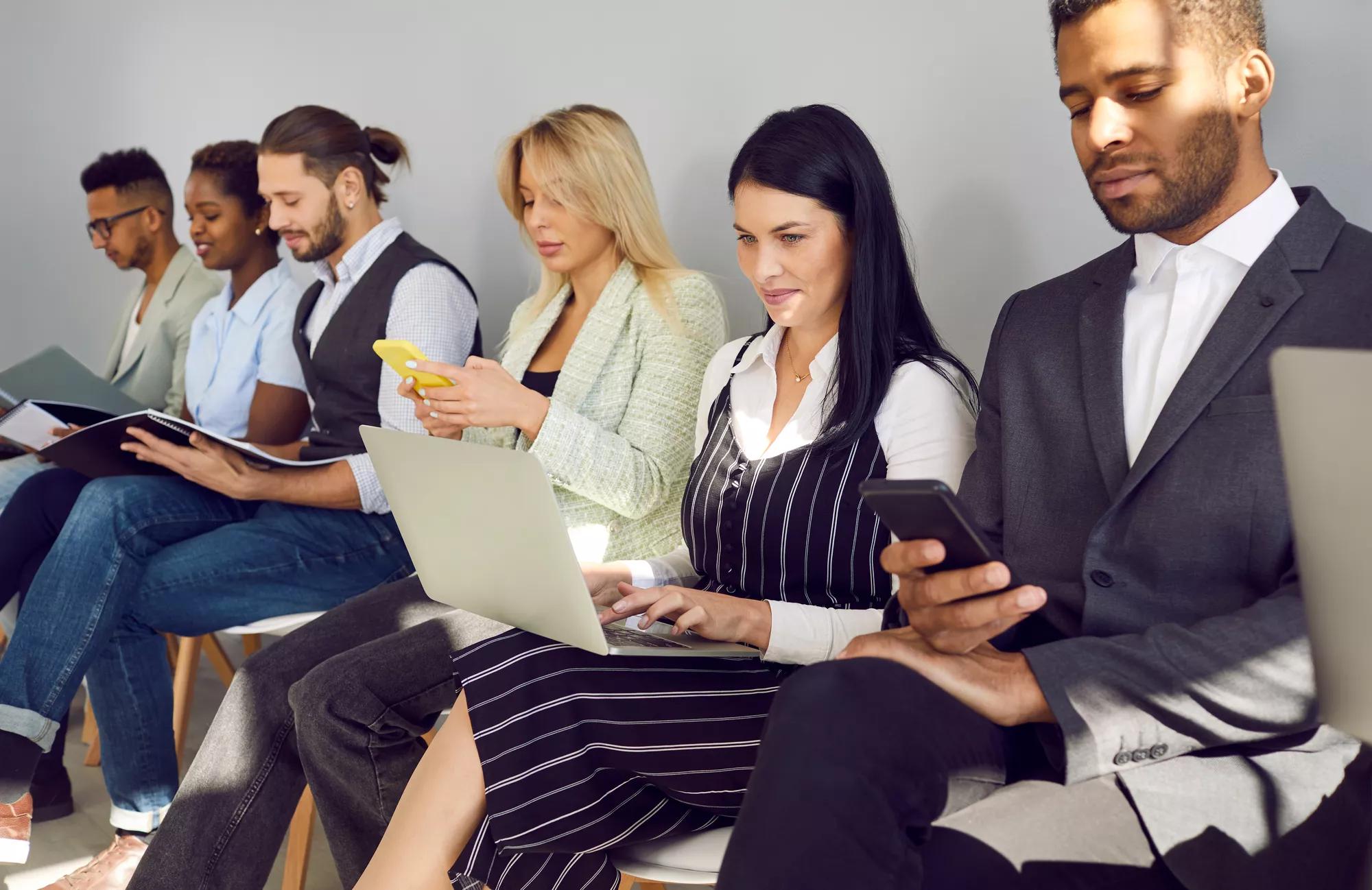Different diverse multiethnic mixed race business people in smart casual clothes holding laptops, using cellphones, reading papers, documents, CVs and resumes while waiting in line for job interview