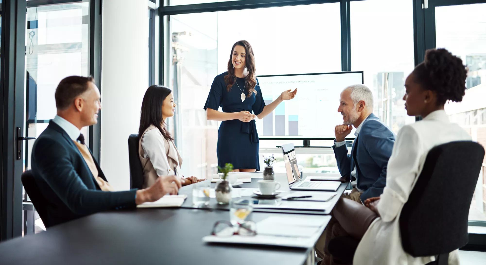 Business woman speaker in an investment meeting with financial data on a boardroom screen. International executive team listening to a presentation. Finance worker talking at a partnership proposal