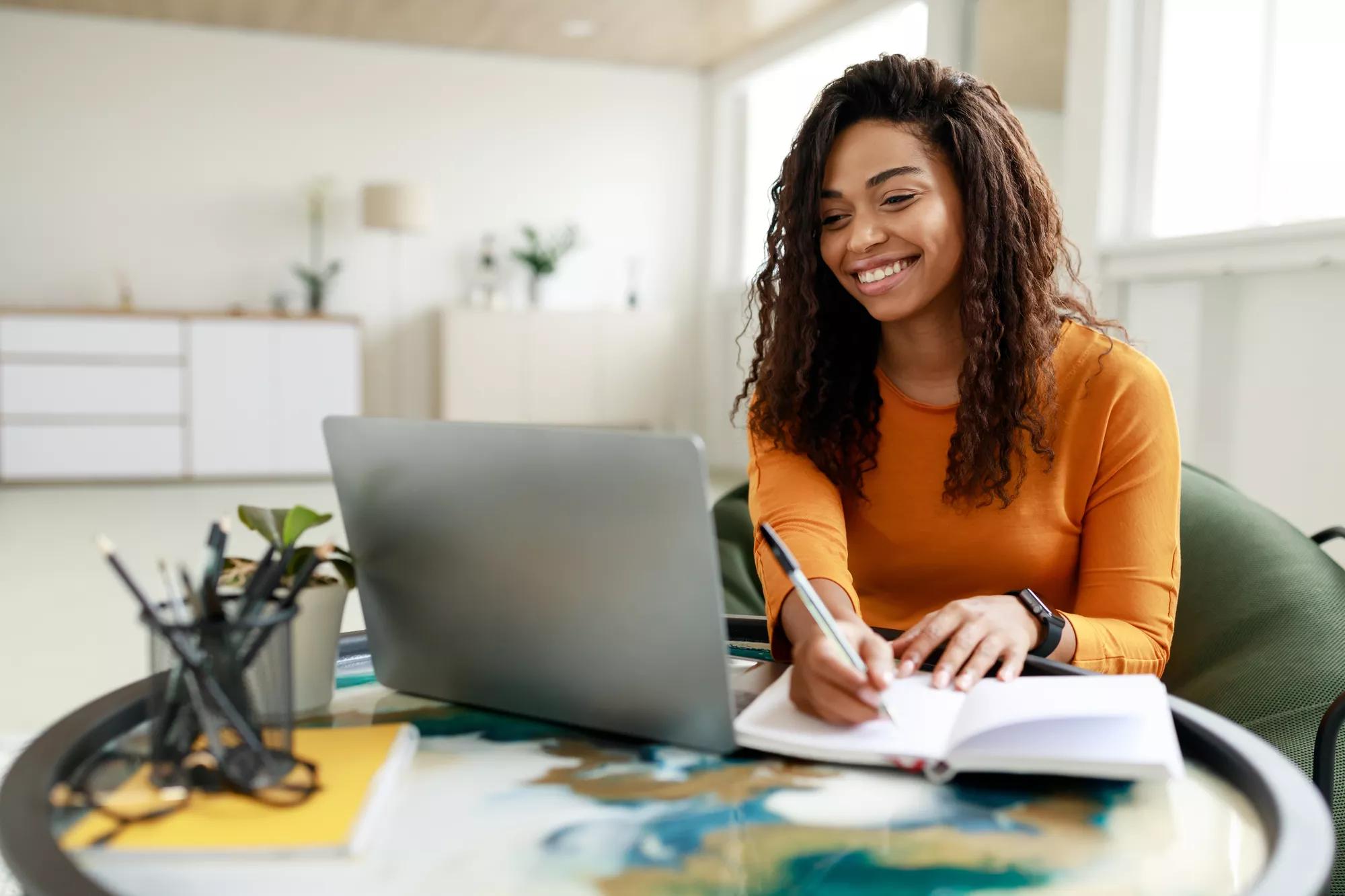 Black woman sitting at desk, using pc writing in notebook