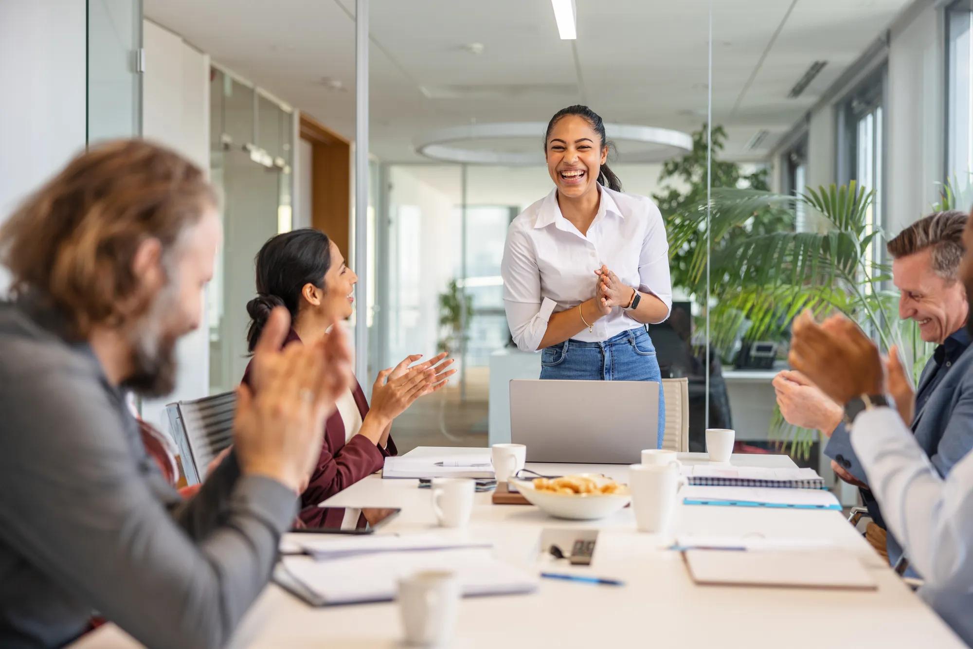 Shot with blur of coworkers from a diverse and multicultural office applauding the great performance of their young dark-skinned female colleague as she is delighted by the praise and smiling back to them.