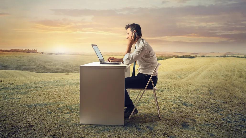 A businessperson wearing a tie sits at a desk. His left hand holds a phone to his ear and his right hand types on a laptop sitting on the desk in front of him. The man, his chair, and desk are all in a wide, expansive field with trees and a sunrise in the distance.