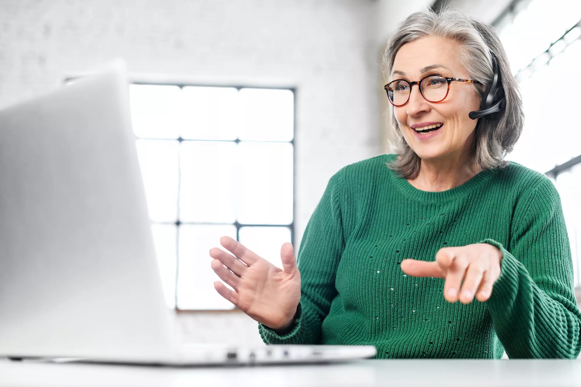 Active mature woman using a laptop for remote work from the home office. Smiling elderly lady wearing wireless headset talking on a video conference, has a video meeting. Senior teacher leads webinar