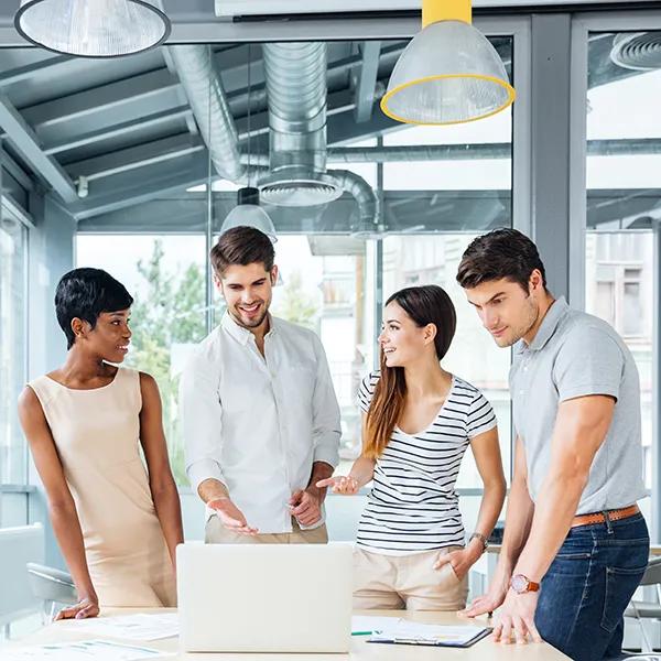 A group of office colleagues stand around a desk looking at a laptop together.