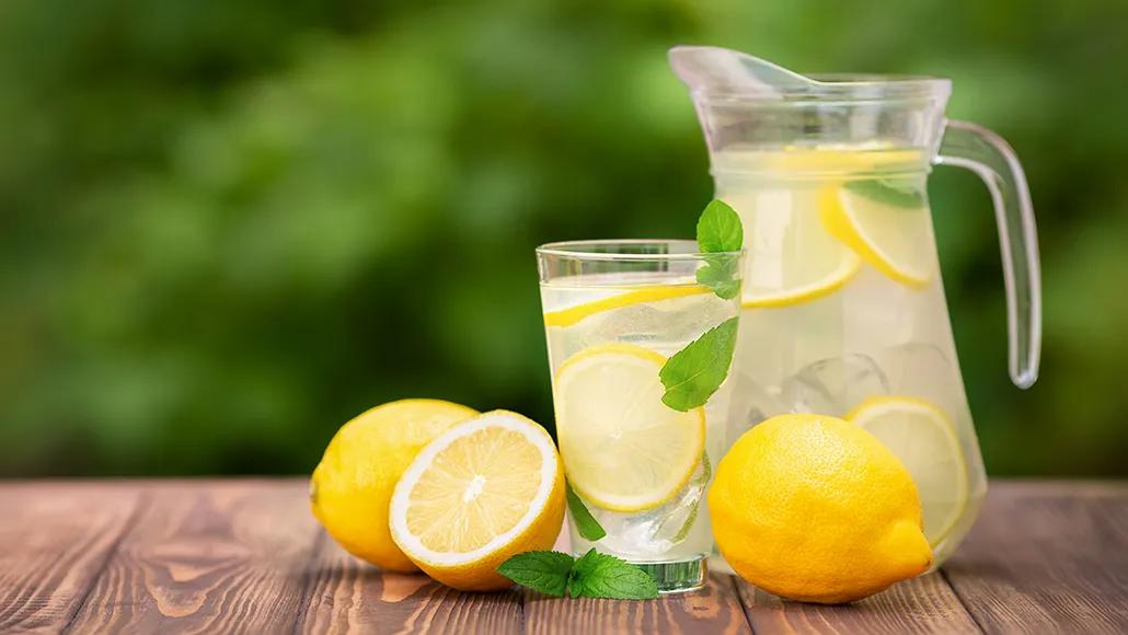 A jug of lemonade, a glass of lemonade, two halves of a lemon, a whole lemon, and a sprig of mint on a wooded table with greenery in the background.