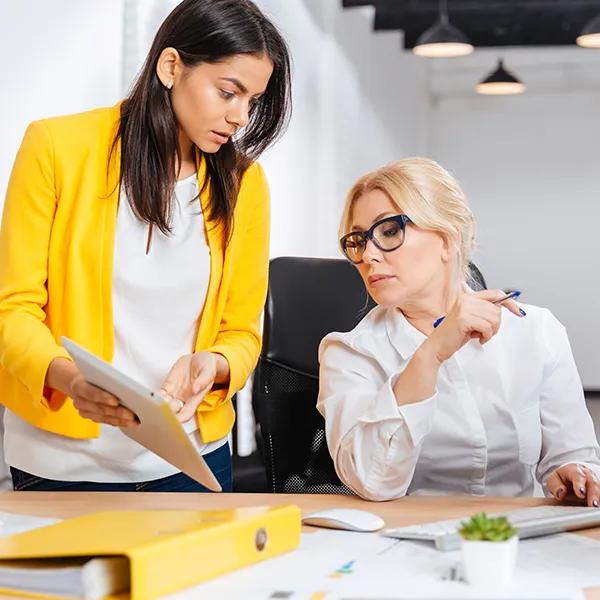 A female colleague is standing at the desk of another female colleague discussing a document.