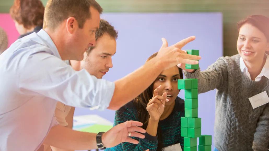 A photo shows colleagues collaboratively stacking green building blocks into a tower.