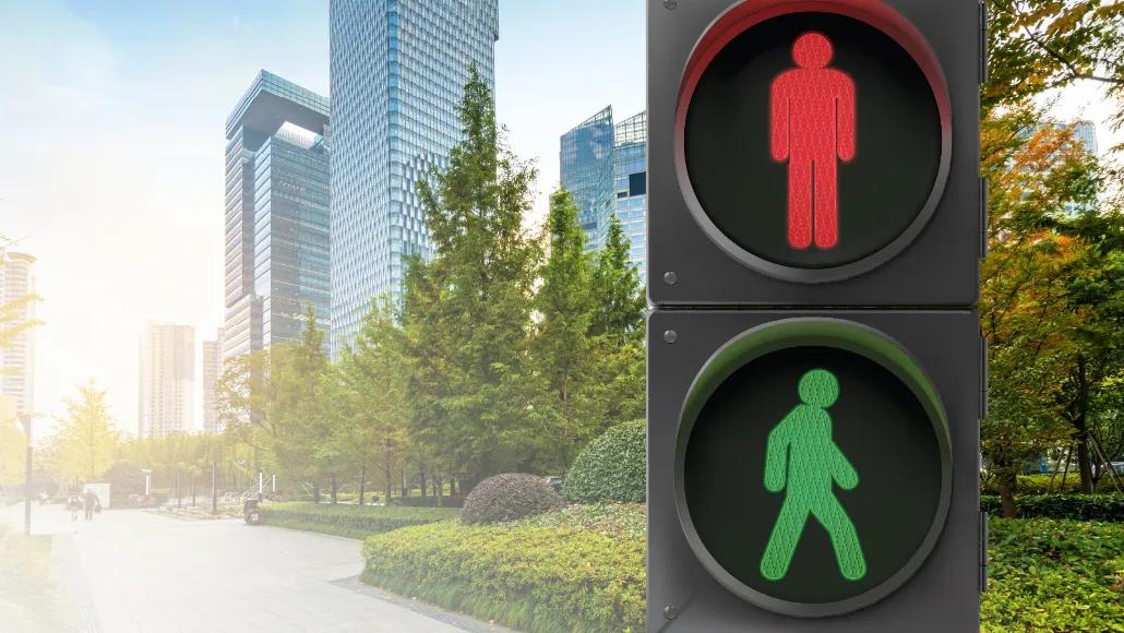 In the background is a bright series of skyscrapers, covered in windows and glass. Next to the skyscrapers is a park with tall trees and manicured bushes. In the foreground is a traffic light. On top of the light is a "stop" icon [a person standing still, arms at sides]. On the bottom of the light is a "go" icon [a person with arms and legs in a walking motion].