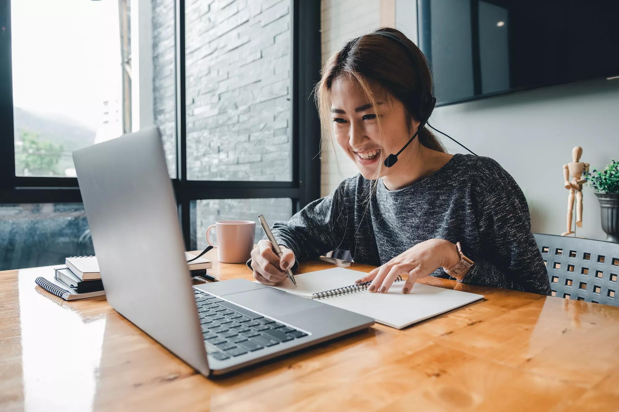 Happy asian woman student watching lesson online and studying from home. Young woman taking notes while looking at computer screen following professor doing math on video call