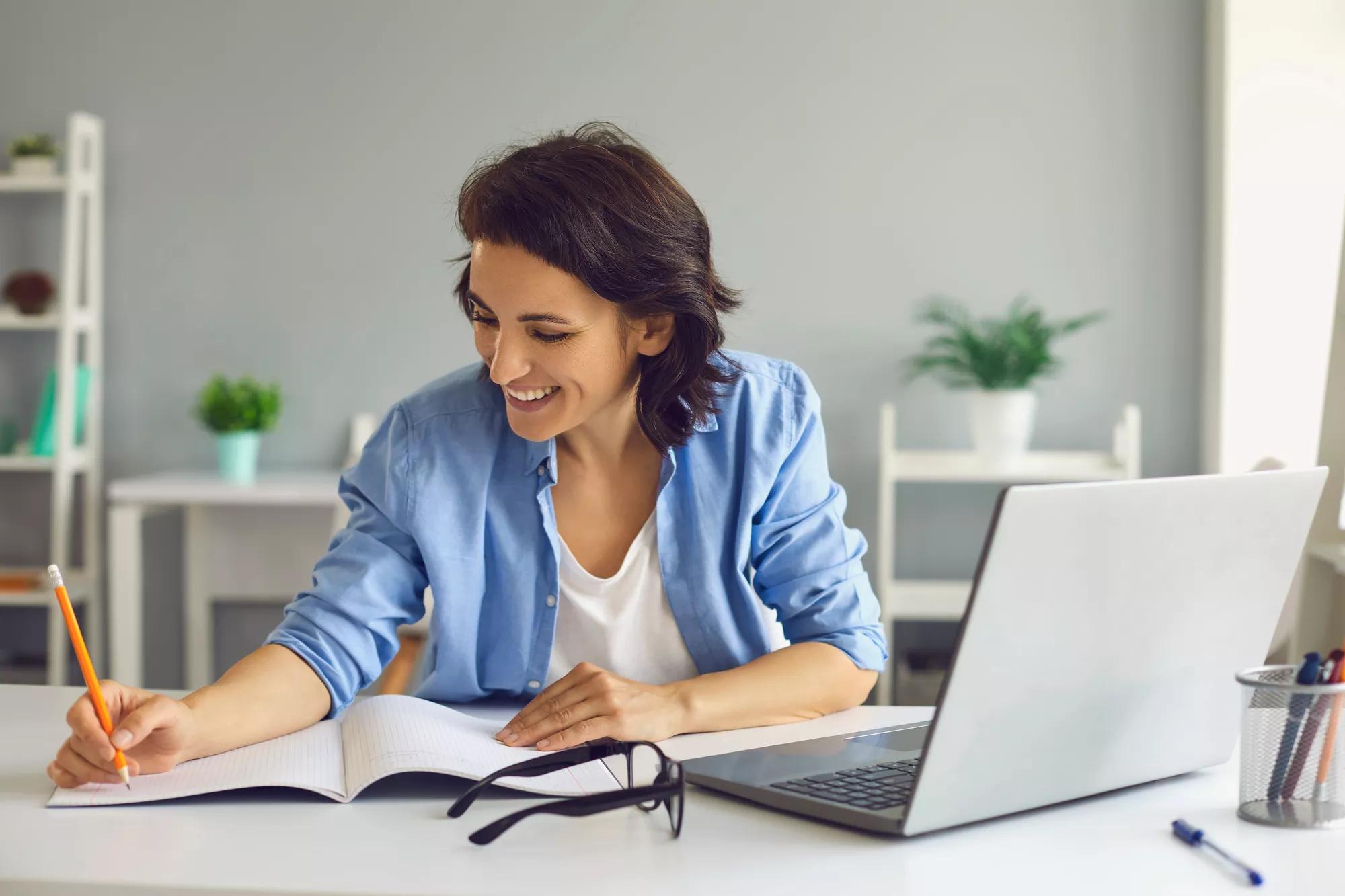 Young smiling woman sitting with laptop and making notes during online conversation, lesson or teleconference with home interior at background. Online communication and elearning concept