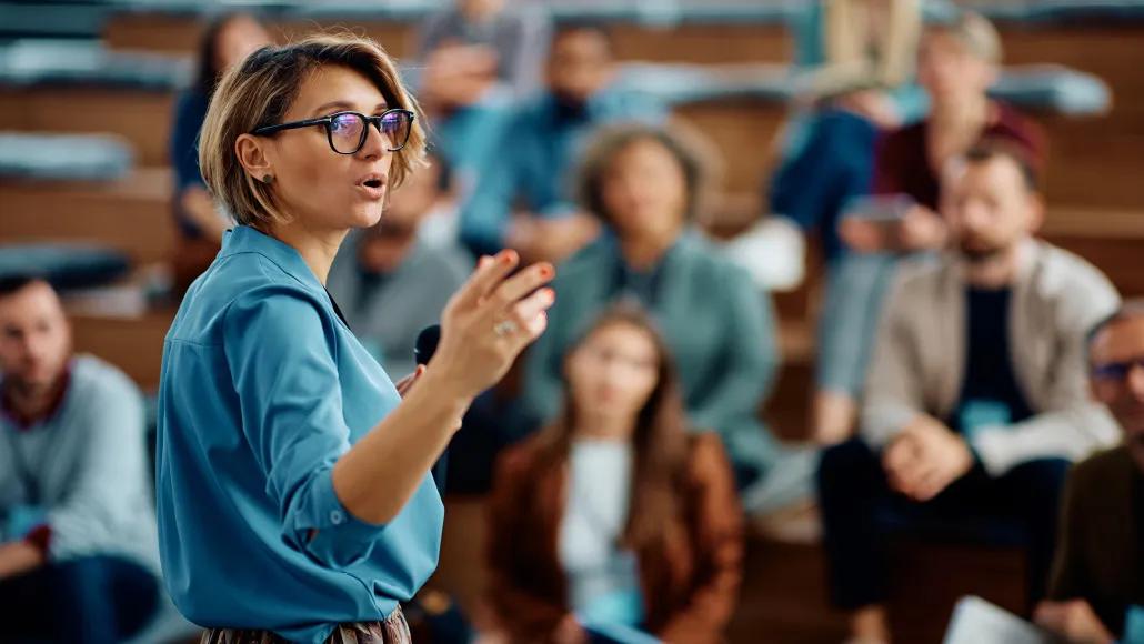 A photograph shows a woman in the foreground with a group of people in the background listening to her speak
