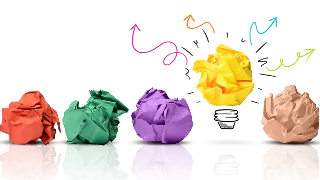 Row of colorful crumpled paper balls one of which is yellow with illustrations making it a lightbulb with arrows radiating outward.